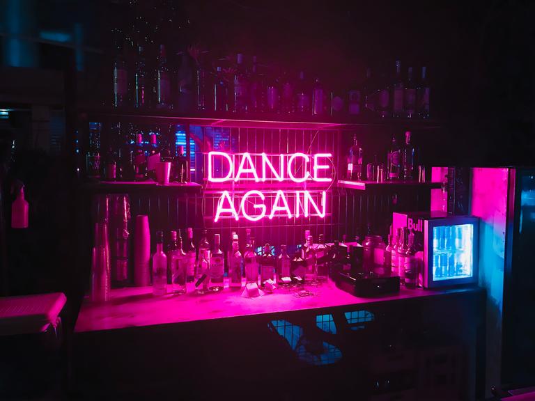 Hinter einer Bar ist in pinkfarbener Neonschrift der Schriftzug "DANCE AGAIN" angebracht.