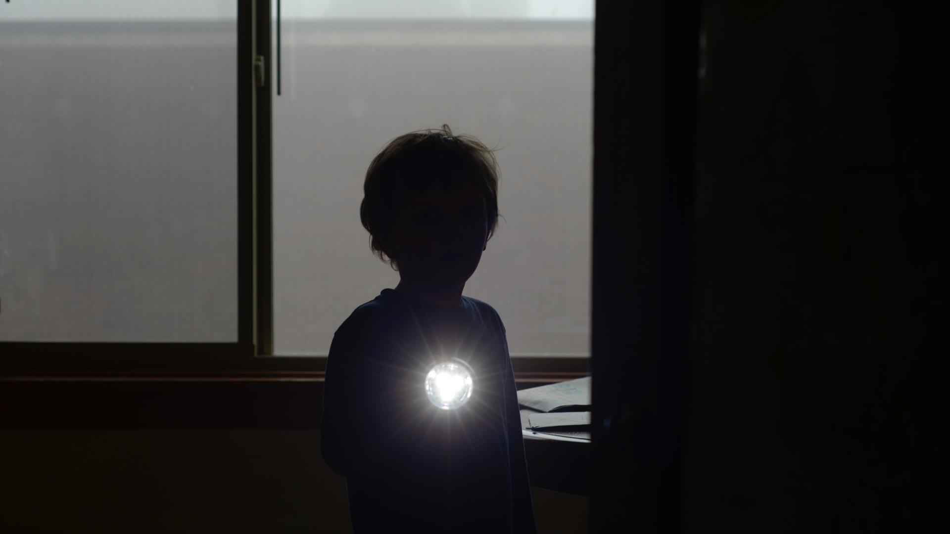 Ein junger Mensch steht mit einer Taschenlampe im Dunkeln und leuchtet in Richtung der Betrachtenden. Zu sehen sind nur Umrisse. Ein junger Mensch steht mit einer Taschenlampe im Dunkeln und leuchtet in Richtung der Betrachtenden. Zu sehen sind nur Umrisse.