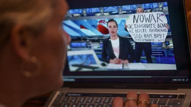 Eine Person schaut sich auf einem Monitor eine Szene aus der abendlichen Hauptnachrichtensendung im russischen Staatsfernsehen an. Darin ist zu sehen, wie eine Kriegsgegnerin die Sendung mit einem Protestplakat unterbricht. 