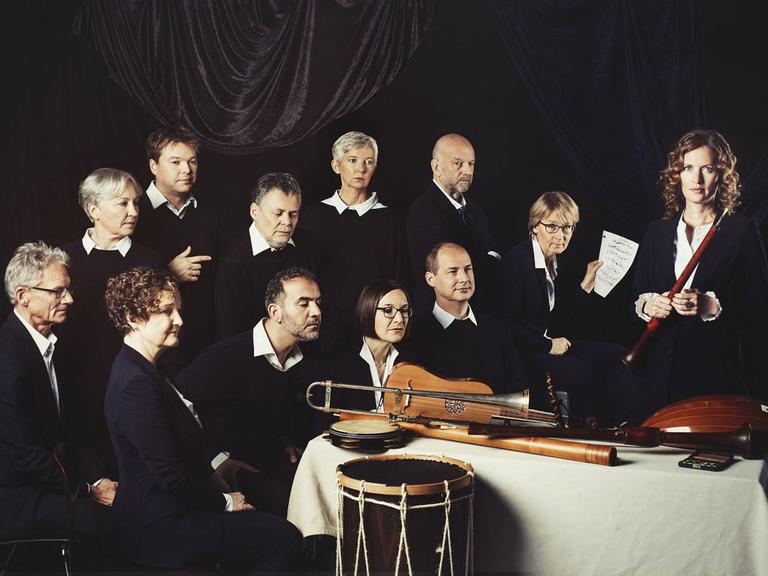 Das Ensemble hat sich in dunklen Kleidern wie auf einem Rembrandgemälde in eine Gruppe um einen Tisch gesetzt, auf dem Musikinstrumente der Renaissance liegen.