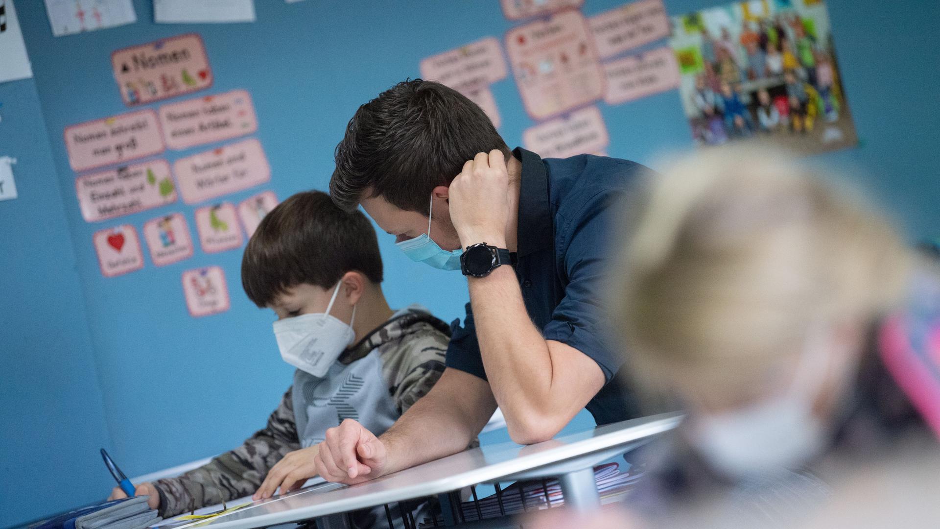 In einem Klassenzimmer sitzt ein Lehrer neben einem kleinen Jungen und schaut ihm bei einer Aufgabe über die Schulter. Beide tragen Mund-Nasenschutz. In einem Klassenzimmer sitzt ein Lehrer neben einem kleinen Jungen und schaut ihm bei einer Aufgabe über die Schulter. Beide tragen Mund-Nasenschutz.