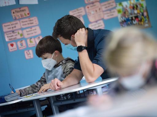 In einem Klassenzimmer sitzt ein Lehrer neben einem kleinen Jungen und schaut ihm bei einer Aufgabe über die Schulter. Beide tragen Mund-Nasenschutz.