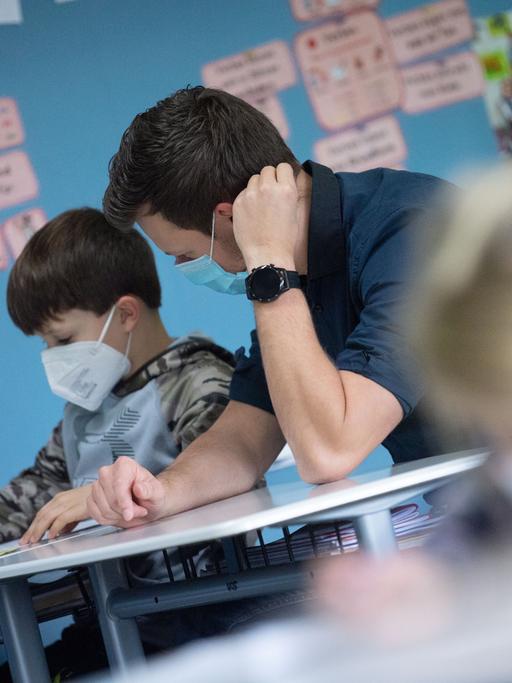 In einem Klassenzimmer sitzt ein Lehrer neben einem kleinen Jungen und schaut ihm bei einer Aufgabe über die Schulter. Beide tragen Mund-Nasenschutz.