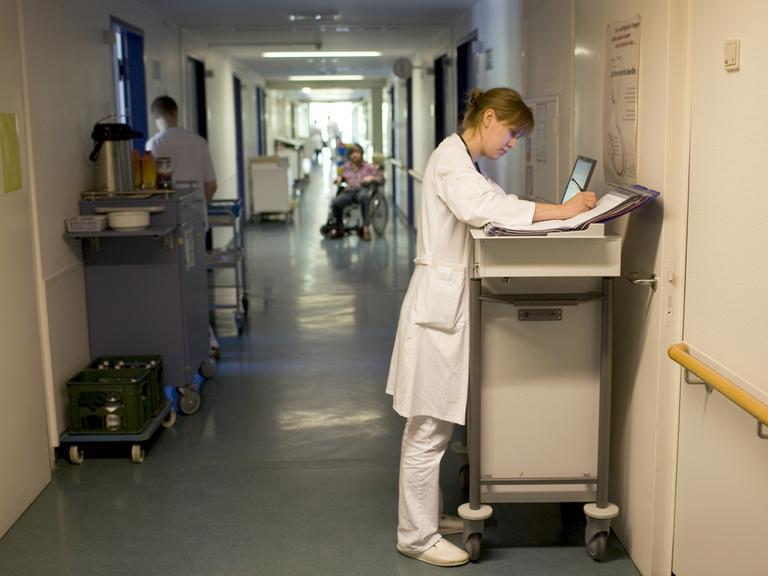 Blick in einen Krankenhausflur, in dem eine Ärztin etwas in ihren Unterlagen notiert. 