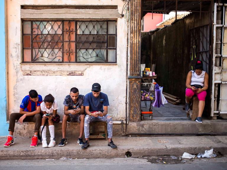 Eine Straßenszene in Kubas Hauptstadt Havana: Fünf Personen, drei Männer, ein Mädchen und eine Frau, sitzen an einem Gehweg an einer Straße und schauen auf ihre Smartphones.