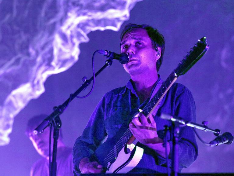 Daniel Rossen steht auf der Bühne am Mikro und spielt Gitarre. Die Beleuchtung taucht den Raum in lila Licht.
