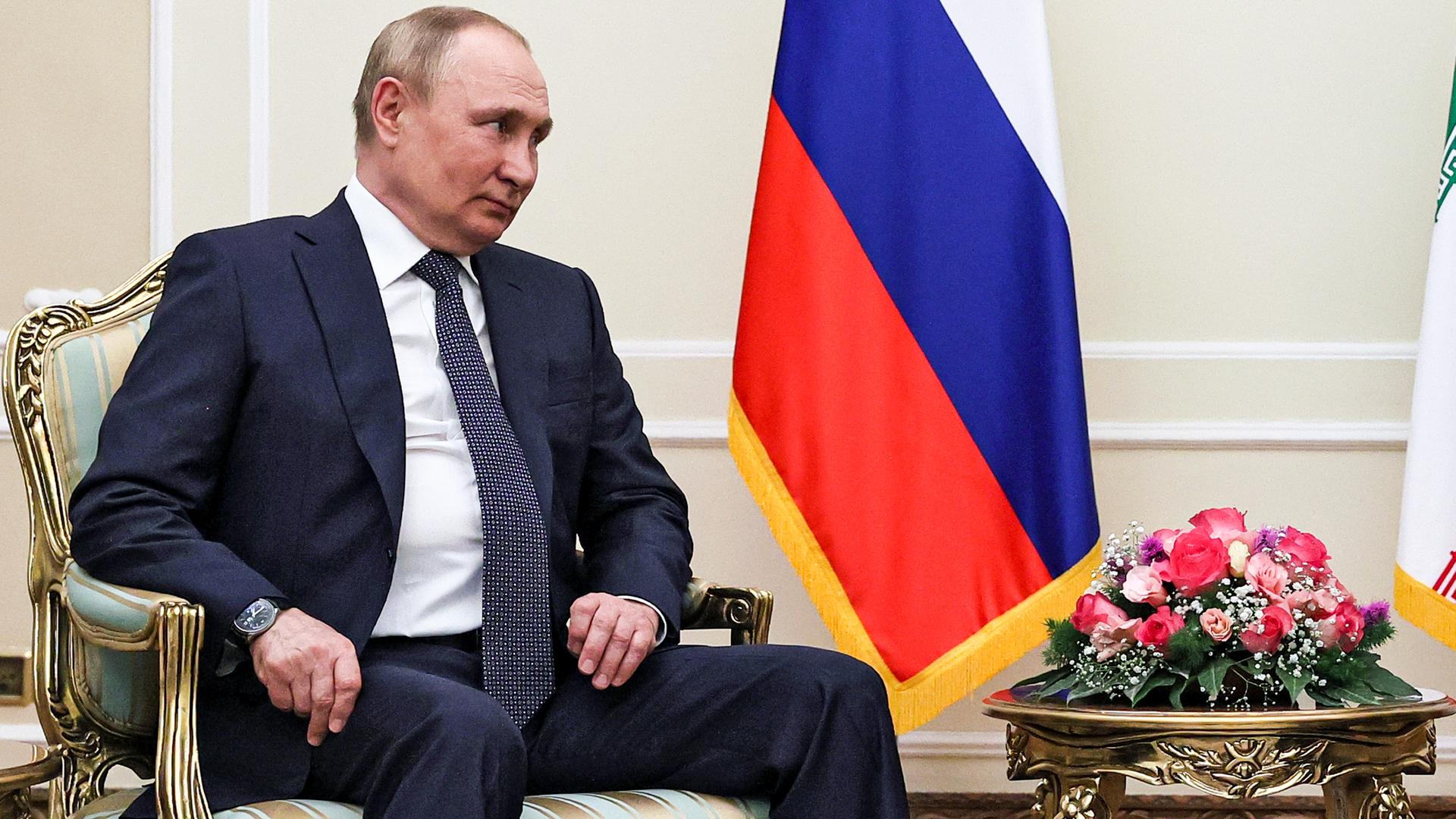 Putin im dunklen Anzug sitzt in einem vergoldeten Sessel neben der weiß-blau-roten Fahne seines Landes und blickt zur Seite. Daneben ein Beistelltisch mit roten Rosen. Putin im dunklen Anzug sitzt in einem vergoldeten Sessel neben der weiß-blau-roten Fahne seines Landes und blickt zur Seite. Daneben ein Beistelltisch mit roten Rosen.