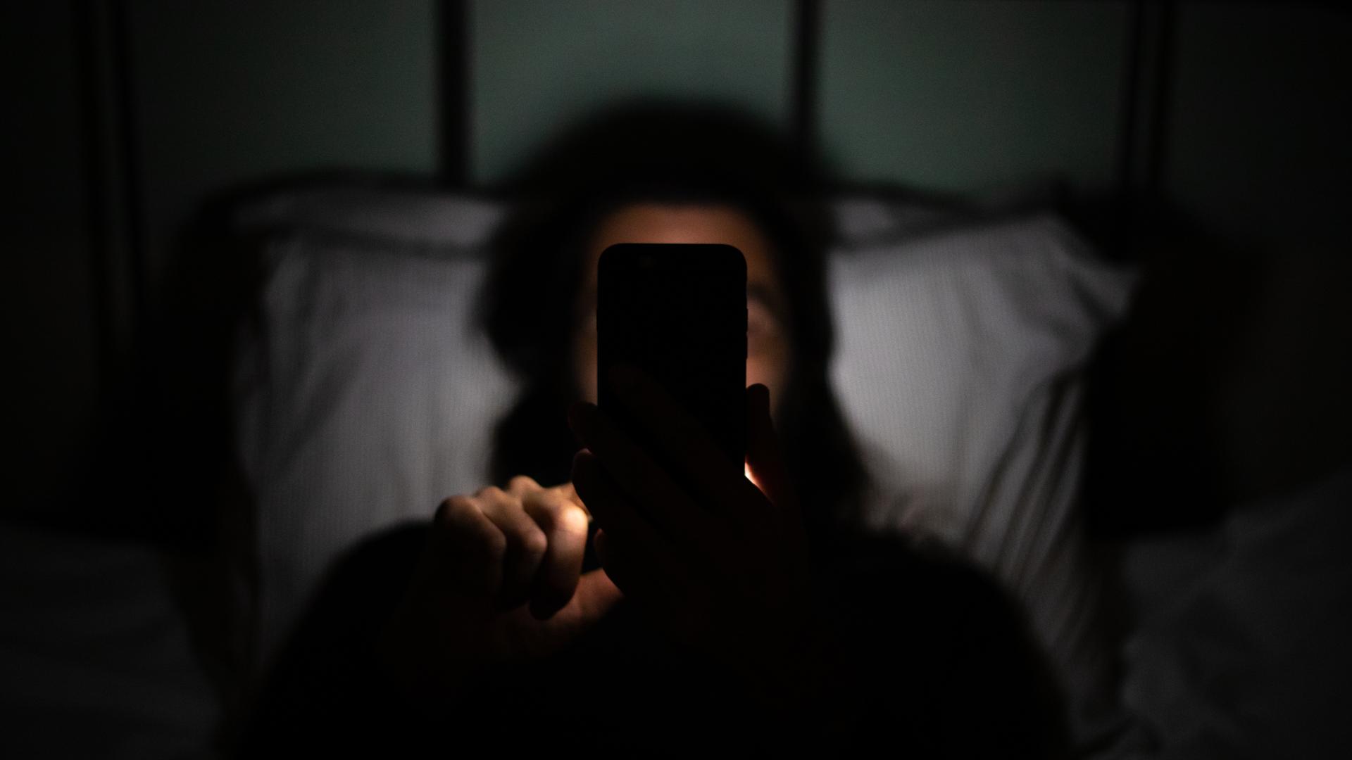 Eine Person liegt im Dunkeln im Bett und hält ihr Smartphone so in der Hand, dass es ihr Gesicht verdeckt.
