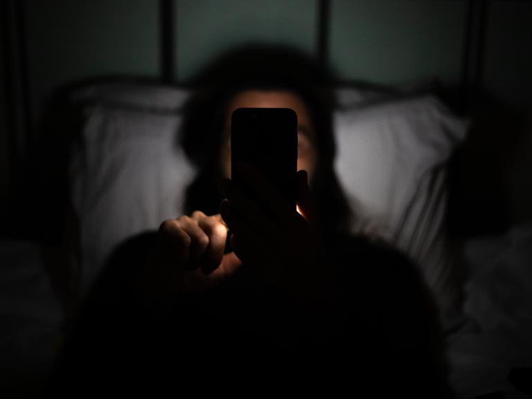 Eine Person liegt im Dunkeln im Bett und hält ihr Smartphone so in der Hand, dass es ihr Gesicht verdeckt.
