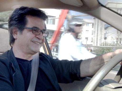 Jafar Panahi sitzt in einem Auto am Steuer und lächelt. 