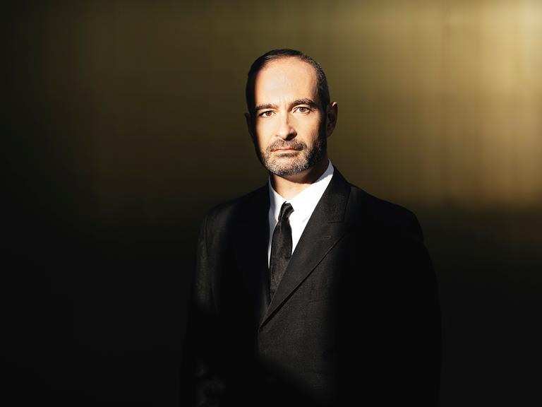 Ein Mann im schwarzen Anzug, kurzen schwarzgrauen Haaren und einem drei Tage Bart blickt in die Kamera. 