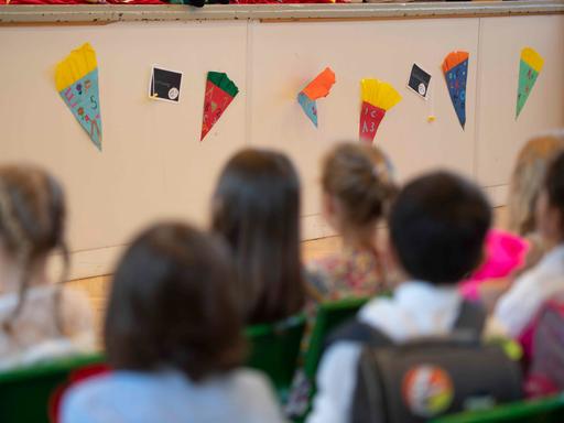 Eine Gruppe Grundschüler ist von hinten zu sehen. Sie sitzen in einer Aula und hören zu.