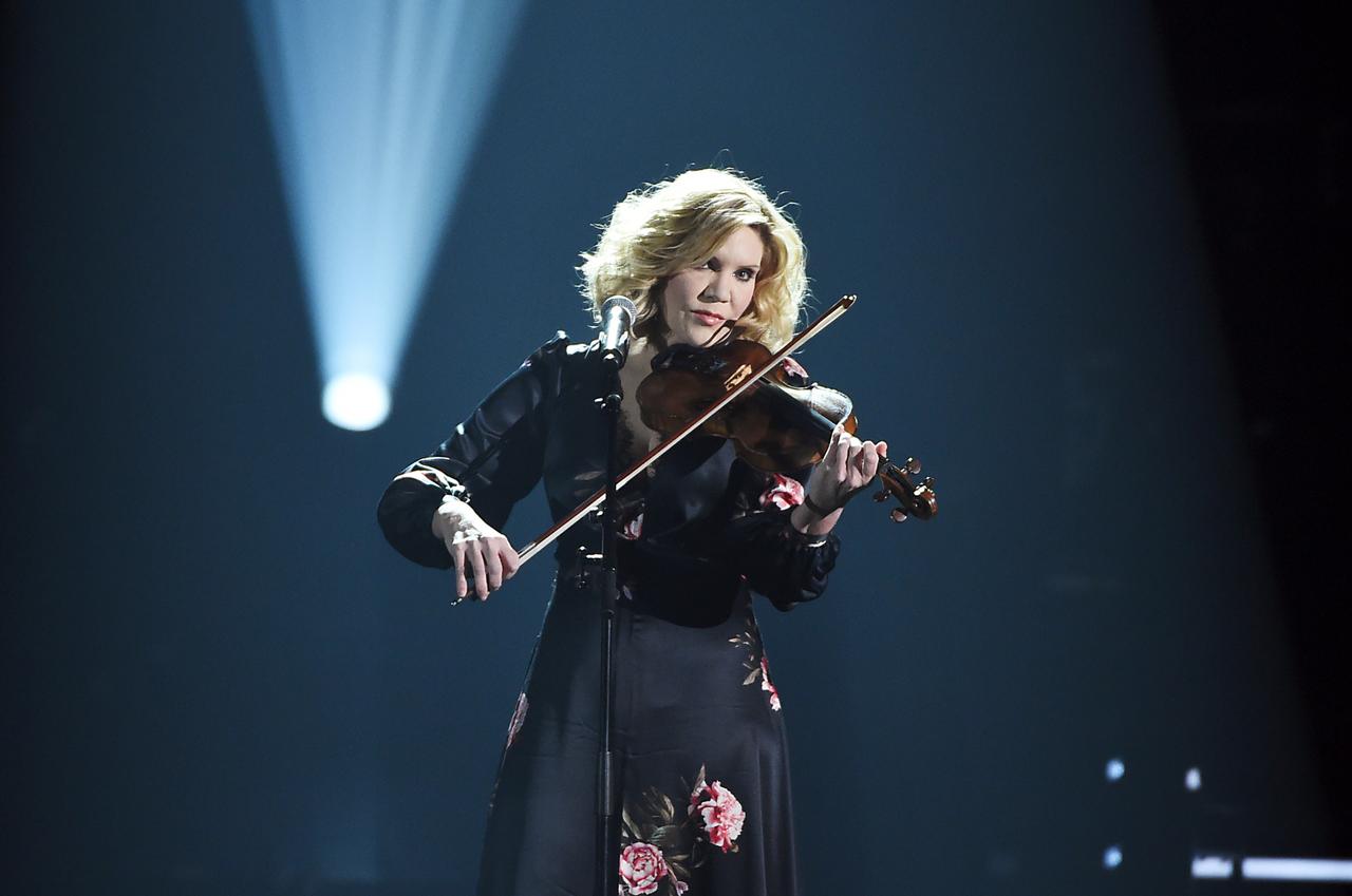 Alison Krauss vor schwarzem Hintergrund auf der Bühne mit einer Geige. Ein Scheinwerfer leuchtet im Hintergrund diffus nach oben.