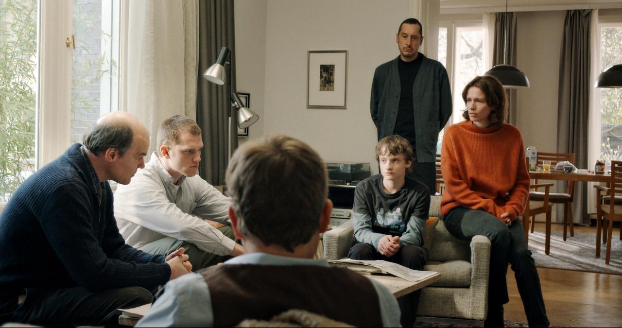 An einem Wohnzimmertisch sitzen Familie und Polizeiermittler beisammen und besprechen die Lage. An einem Wohnzimmertisch sitzen Familie und Polizeiermittler beisammen und besprechen die Lage.