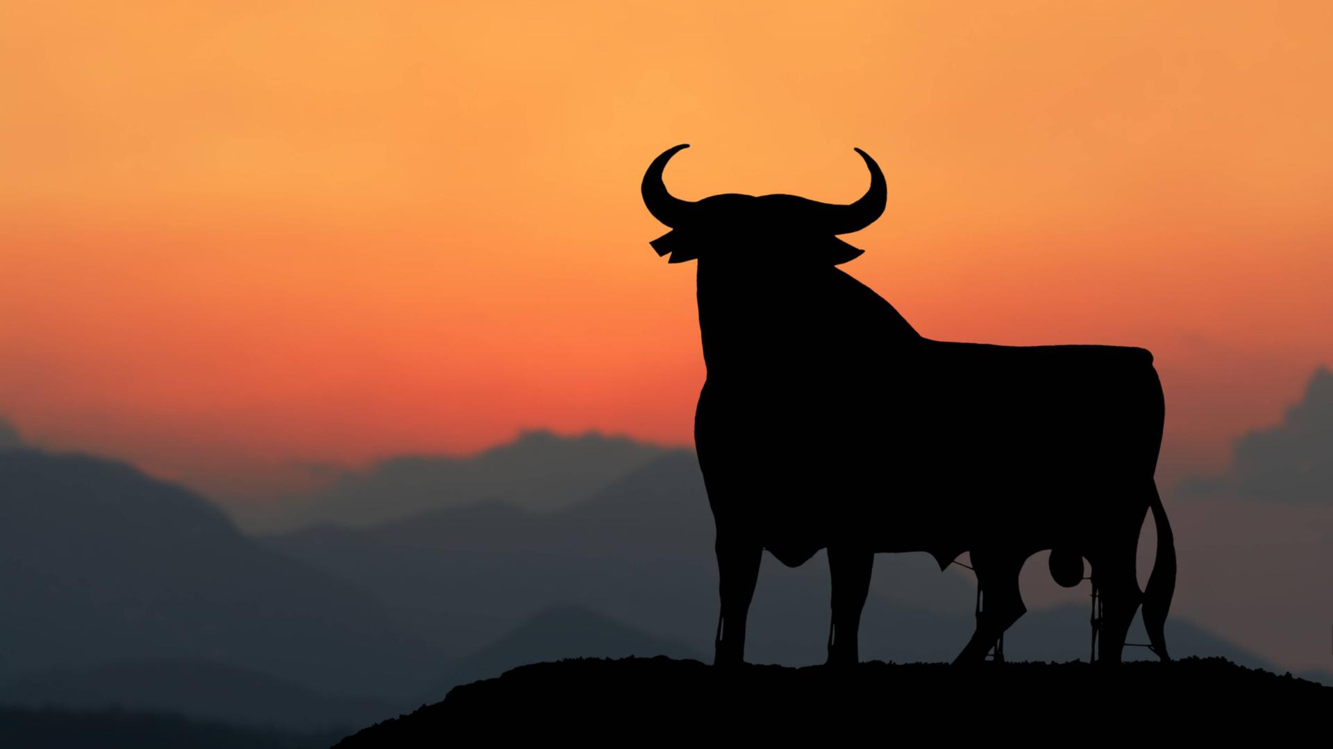 Die Silhouette eines großen schwarzen Stiers mit gerundeten Hörnern steht im Abendrot auf dem Gipfel eines Berges inmitten eines Gebirges. Die Silhouette eines großen schwarzen Stiers mit gerundeten Hörnern steht im Abendrot auf dem Gipfel eines Berges inmitten eines Gebirges.