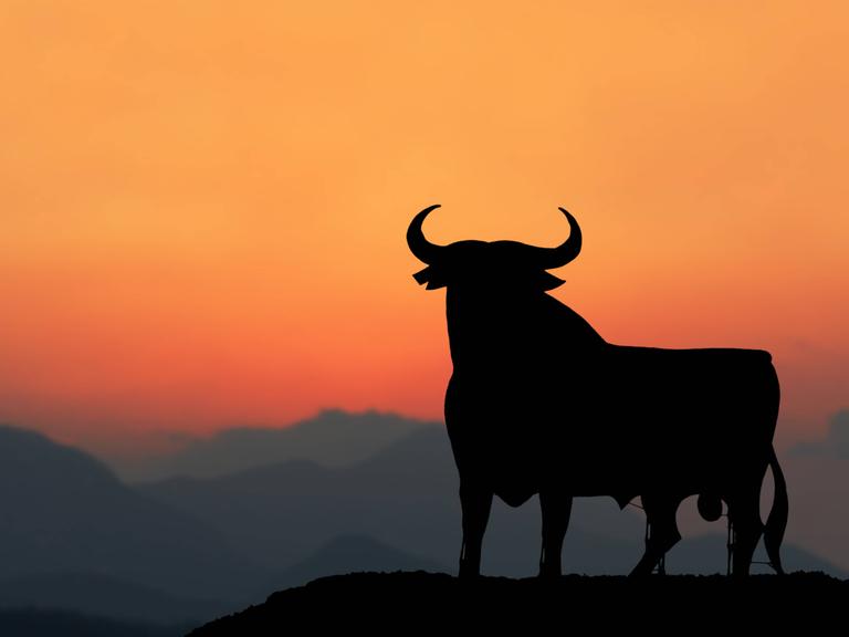 Die Silhouette eines großen schwarzen Stiers mit gerundeten Hörnern steht im Abendrot auf dem Gipfel eines Berges inmitten eines Gebirges.