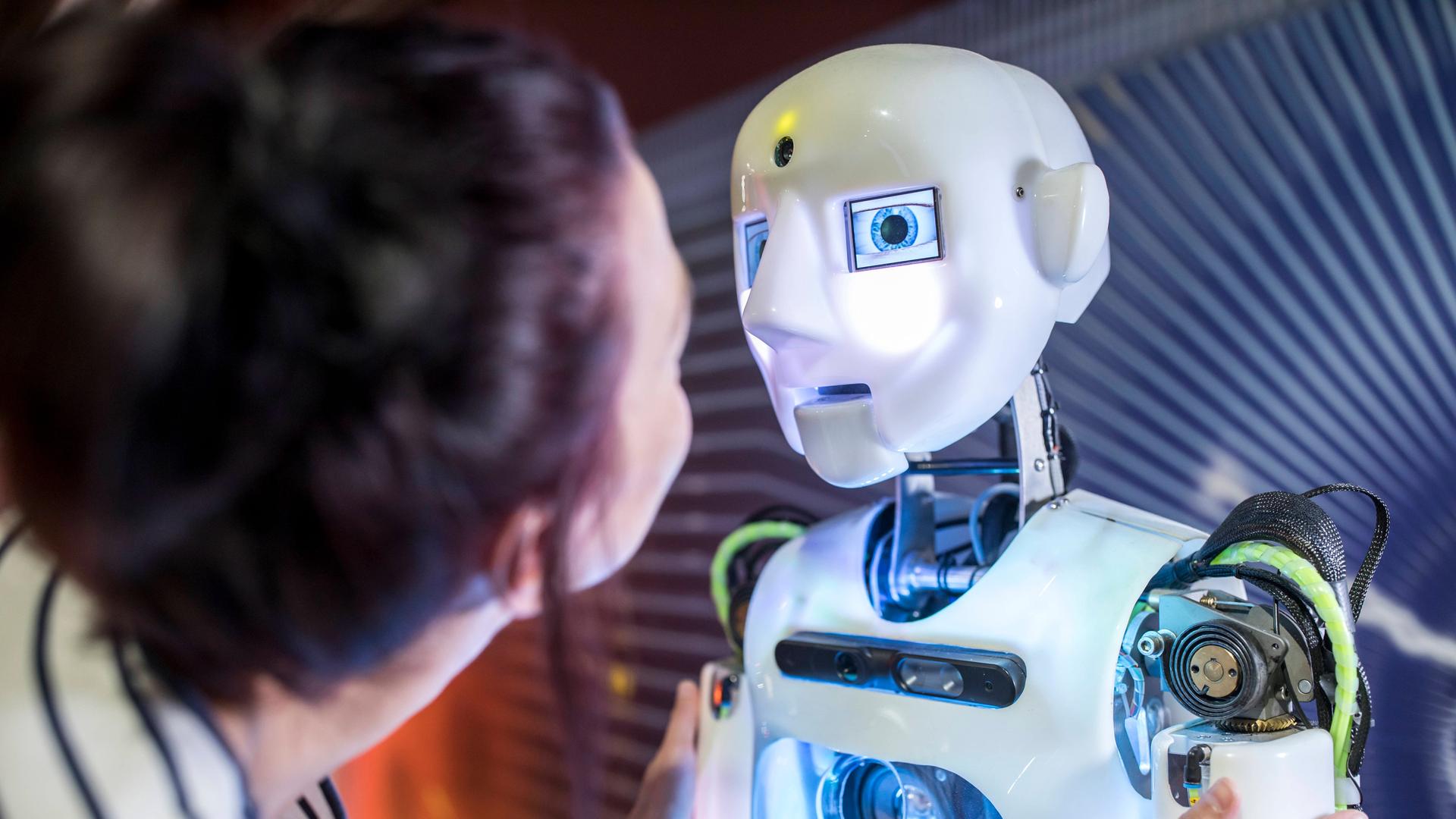 Eine junge Frau steht halb vor einem Roboter mit menschlich nachempfundenem Gesicht, den die Frau am Oberarm berührt und mit ihm zu sprechen scheint. Eine junge Frau steht halb vor einem Roboter mit menschlich nachempfundenem Gesicht, den die Frau am Oberarm berührt und mit ihm zu sprechen scheint.