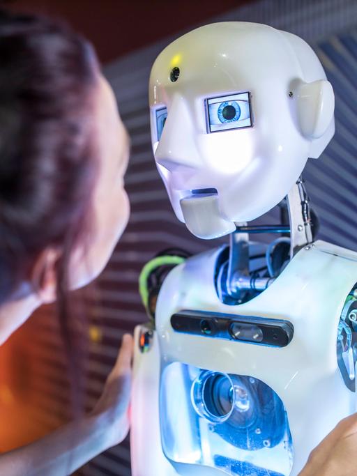 Eine junge Frau steht halb vor einem Roboter mit menschlich nachempfundenem Gesicht, den die Frau am Oberarm berührt und mit ihm zu sprechen scheint.
