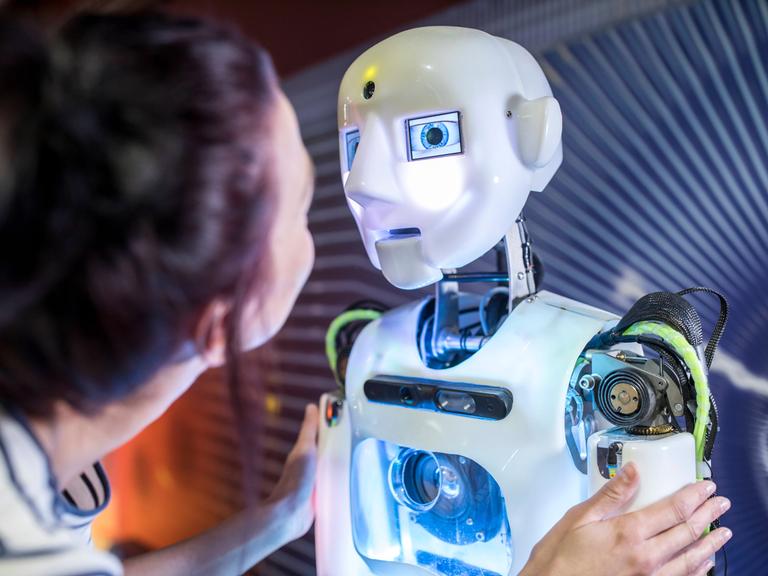 Eine junge Frau steht halb vor einem Roboter mit menschlich nachempfundenem Gesicht, den die Frau am Oberarm berührt und mit ihm zu sprechen scheint.