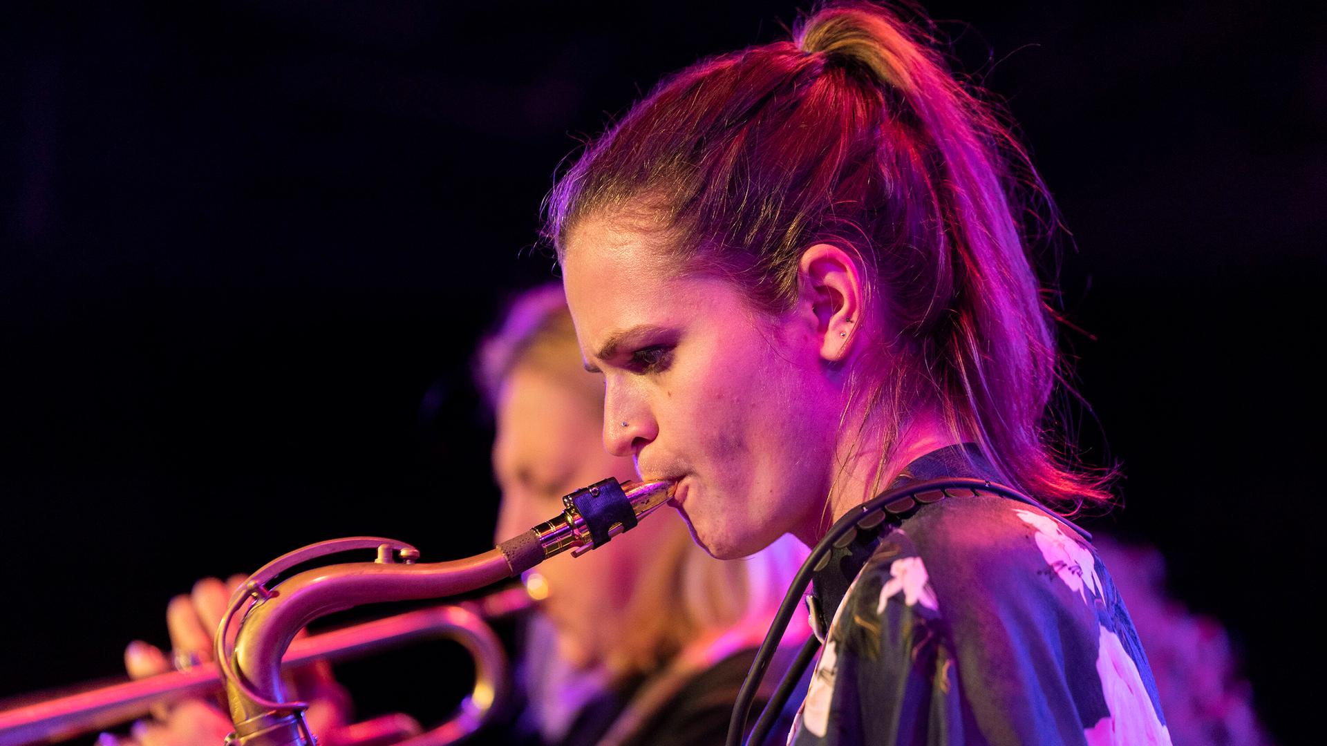 Nicole Glover steht mit brünettem Pferdeschwanz vor einem Musikerkollegen auf einer lila erleuchteten Bühne und hat das Tenorsaxophon an die Lippen angesetzt. Nicole Glover steht mit brünettem Pferdeschwanz vor einem Musikerkollegen auf einer lila erleuchteten Bühne und hat das Tenorsaxophon an die Lippen angesetzt.