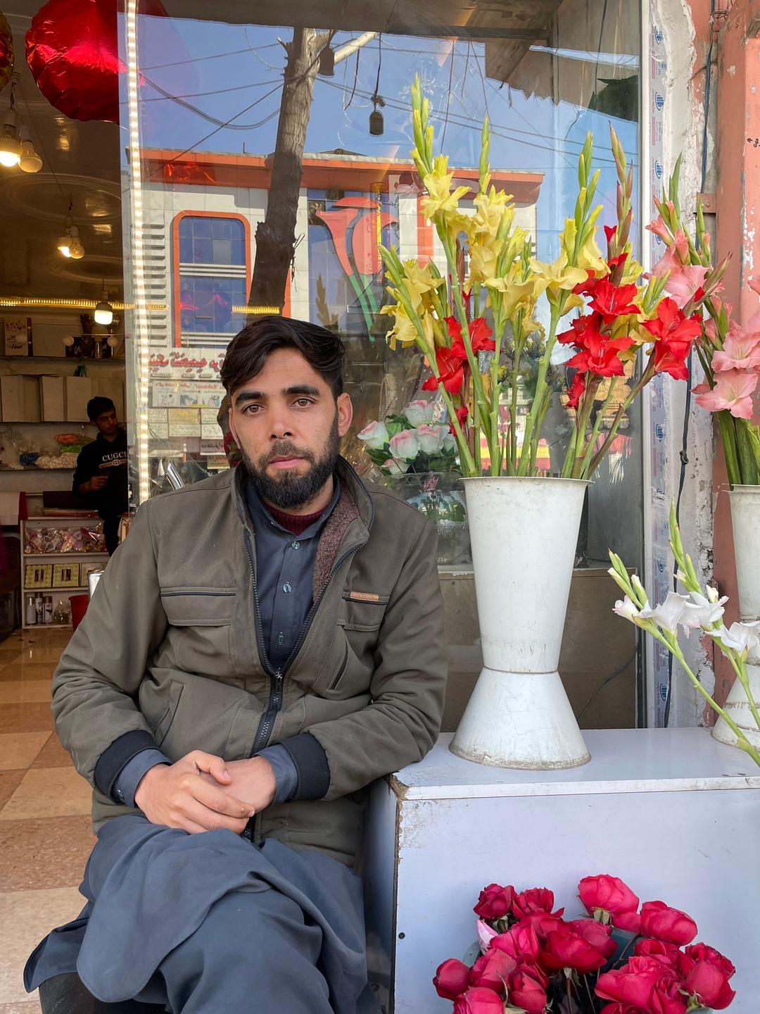 Ein junger Mann mit Vollbart sitzt vor dem Laden auf der Straße neben Sträußen von bunten Blumen in Vasen.