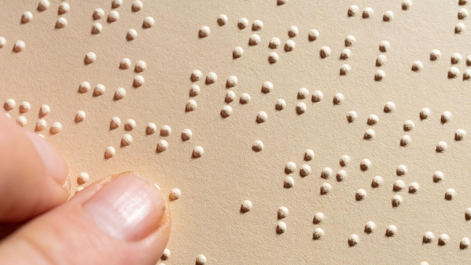 Ein Finger auf beigefarbenem Untergrund. Davon heben sich die Punkte der Brailleschrift ab. Ein Finger auf beigefarbenem Untergrund. Davon heben sich die Punkte der Brailleschrift ab.