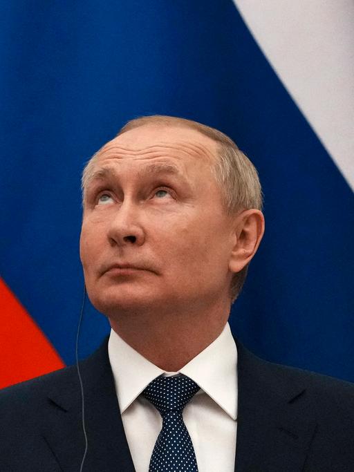 Wladimir Putin mit nach oben gerichteten Blick. Im Hintergrund ist die russiche Flagge zu sehen.