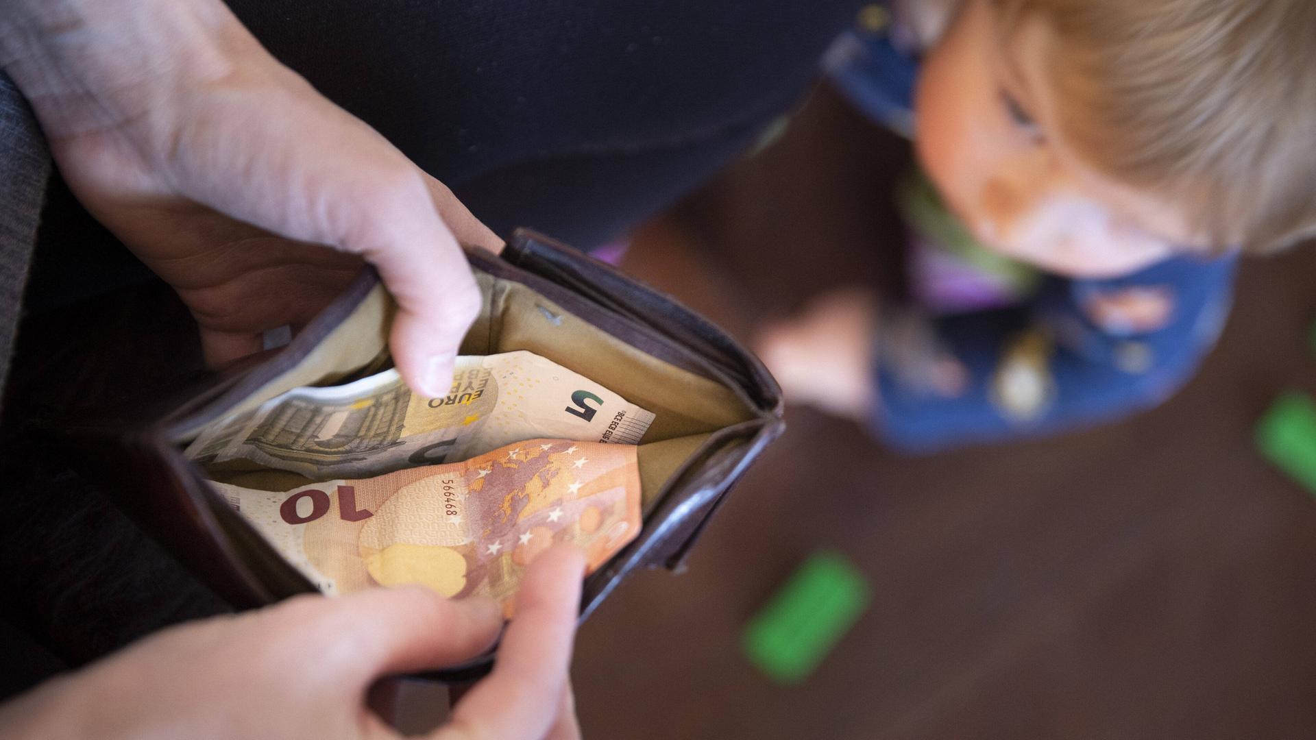 Blick von oben auf eine geöffnete Geldbörse und ein Kind, das auf diese schaut. In der Geldböse ist ein fünf und ein zehn Euro-Schein. Blick von oben auf eine geöffnete Geldbörse und ein Kind, das auf diese schaut. In der Geldböse ist ein fünf und ein zehn Euro-Schein.
