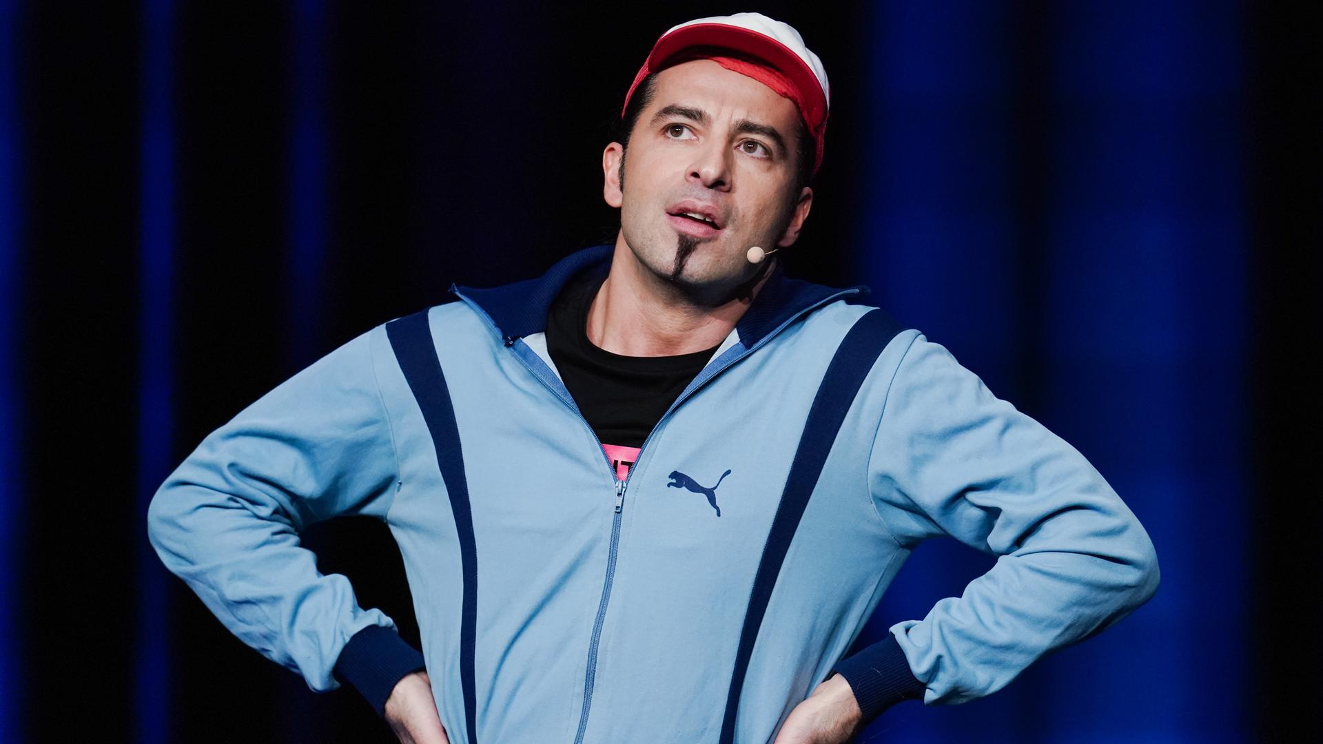 Bülent Ceylan steht mit blauer Trainingsjacke und rot-weißer Cappy auf einer Bühne und guckt leicht fragend nach oben. Bülent Ceylan steht mit blauer Trainingsjacke und rot-weißer Cappy auf einer Bühne und guckt leicht fragend nach oben.