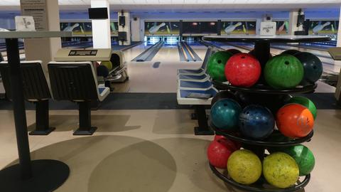 Im Vordergrund ist ein Halter für Bowlingkugeln, weiter hinten ein etwas in die Jahre gekommenes Bowlingcenter mit Bahnen und Punktezählern.