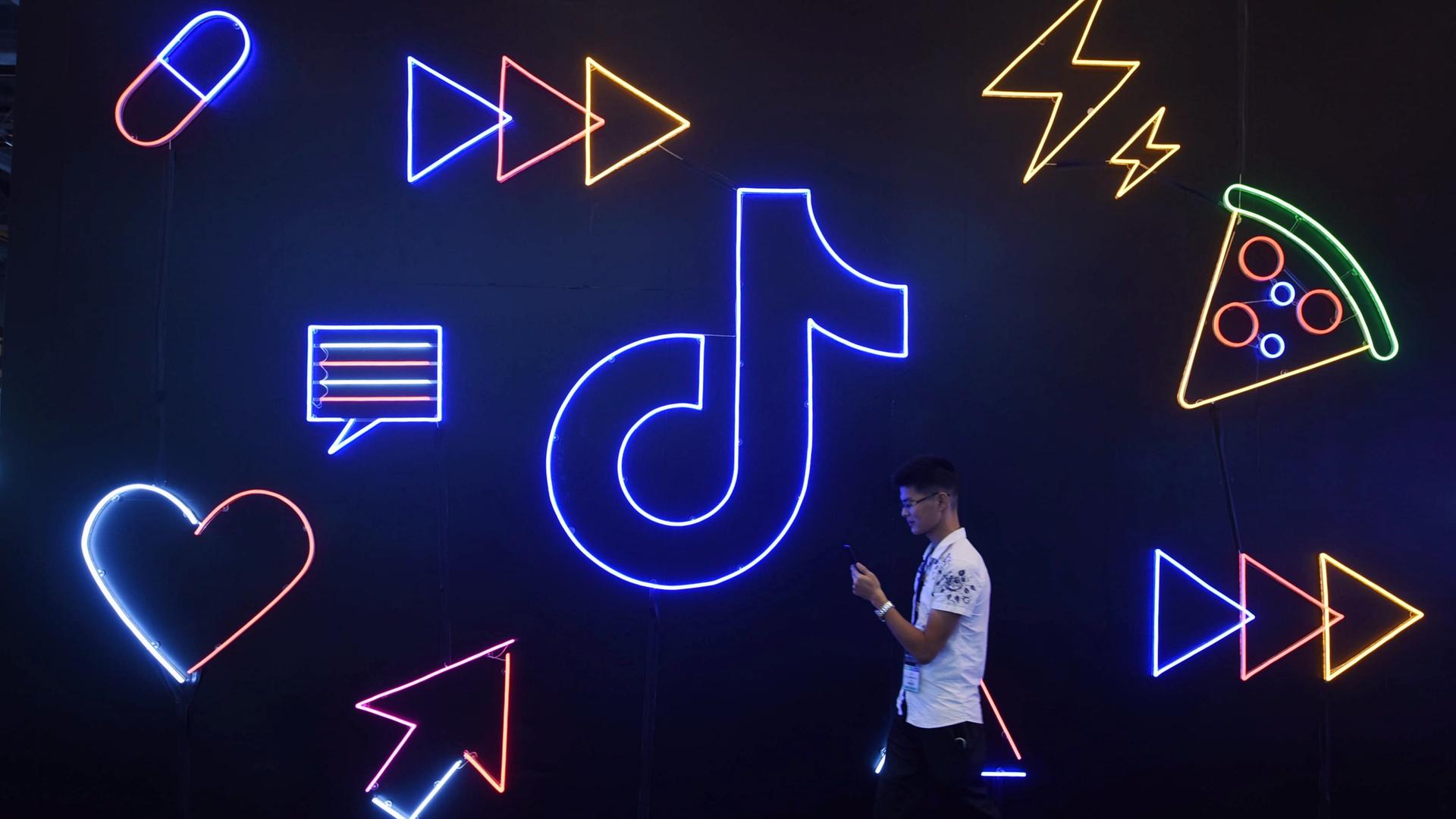 Ein Mann läuft mit seinem Smartphone vor einer Wand entlang, an der in Neonfarben das TikTok-Logo, Herzen und Pfeile prangen. Ein Mann läuft mit seinem Smartphone vor einer Wand entlang, an der in Neonfarben das TikTok-Logo, Herzen und Pfeile prangen.