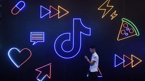 Ein Mann läuft mit seinem Smartphone vor einer Wand entlang, an der in Neonfarben das TikTok-Logo, Herzen und Pfeile prangen.