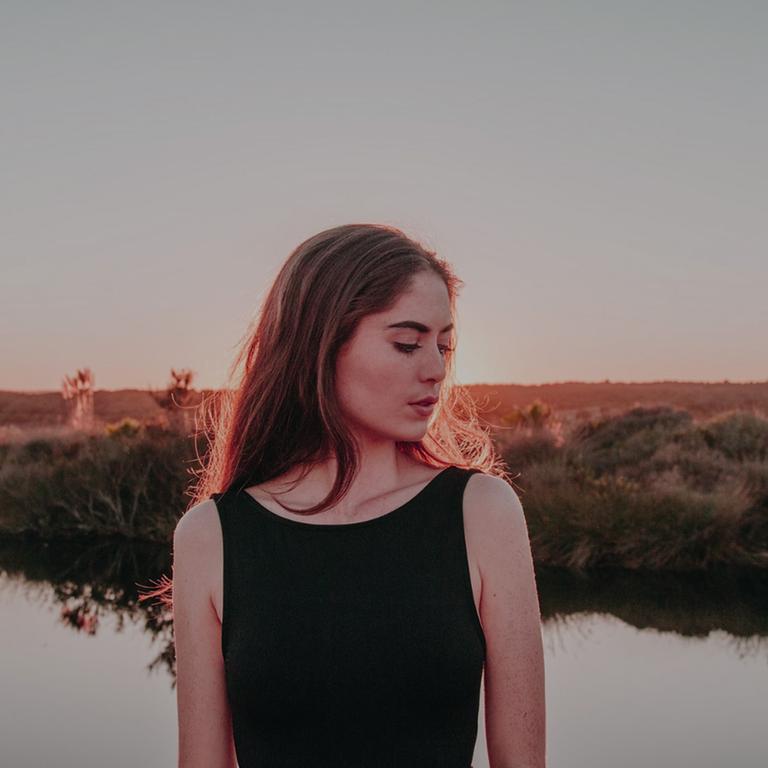 Eine junge Frau, mit dem Kopf zur Seite, steht vor einem See. Im Hintergrund ist eine flache Landschaft zu sehen. Eine junge Frau, mit dem Kopf zur Seite, steht vor einem See. Im Hintergrund ist eine flache Landschaft zu sehen.
