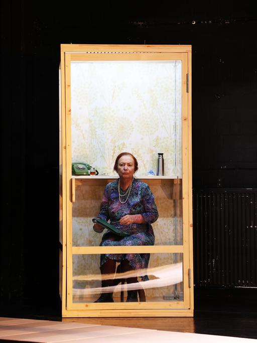 Gabriele Möller-Lukasz sitzt in der Inszenierung von "Mutter Vater Land" auf einem Stuhl hinter einer Glastür.