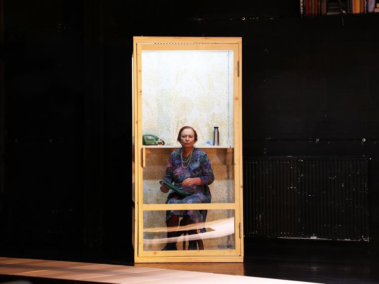 Gabriele Möller-Lukasz sitzt in der Inszenierung von "Mutter Vater Land" auf einem Stuhl hinter einer Glastür.