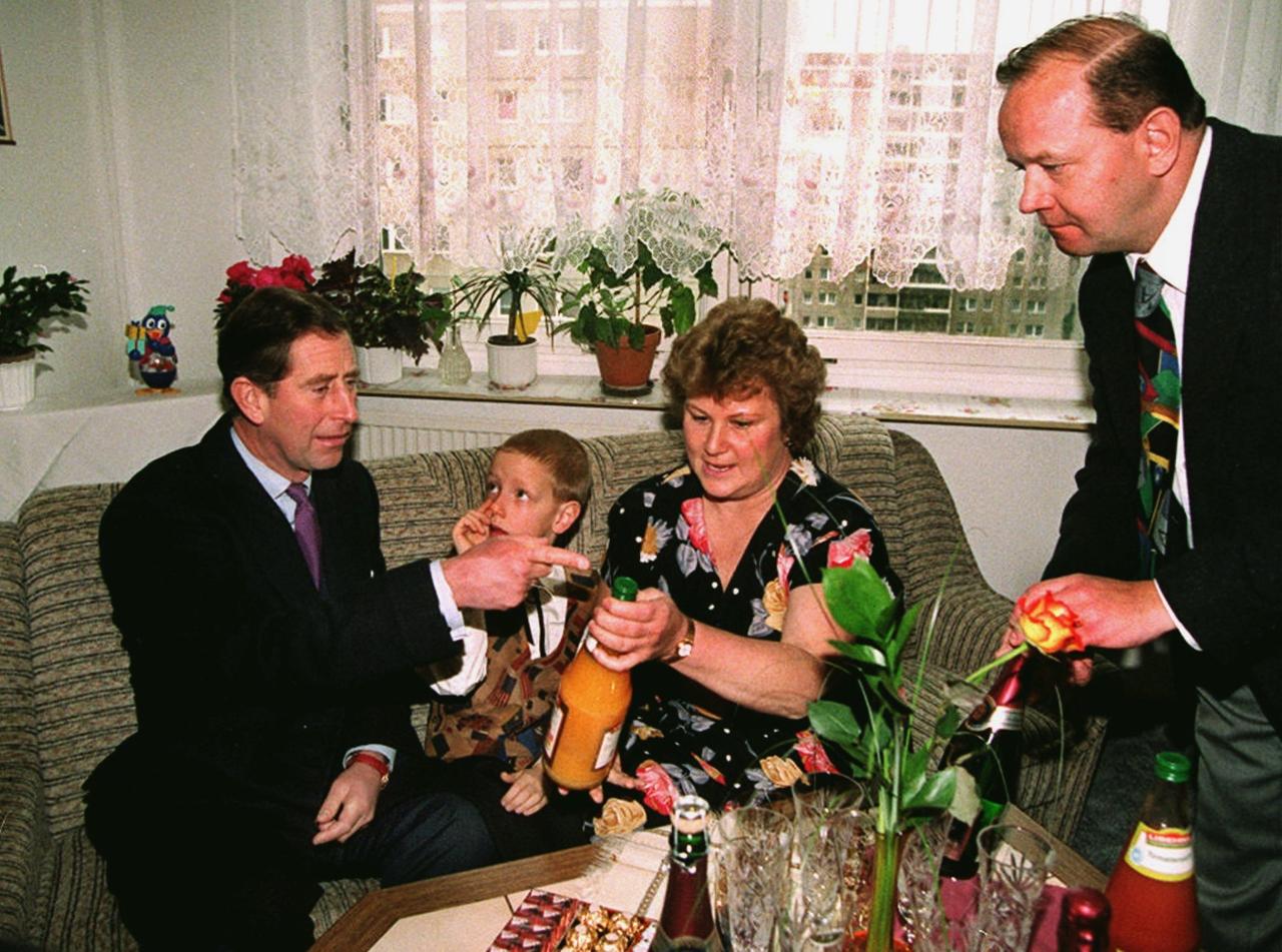 Prinz Charles sitzt mit einer Familie auf der Couch im Wohzimmer einer Plattenbau-Wohnung. Prinz Charles sitzt mit einer Familie auf der Couch im Wohzimmer einer Plattenbau-Wohnung.