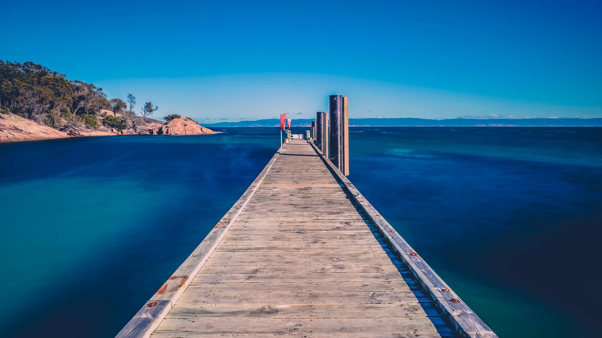 Ein lang gezogener Anlegesteg aus Holz führt unter einem knallblauen Himmel durch tiefblaues Wasser. Ein lang gezogener Anlegesteg aus Holz führt unter einem knallblauen Himmel durch tiefblaues Wasser.