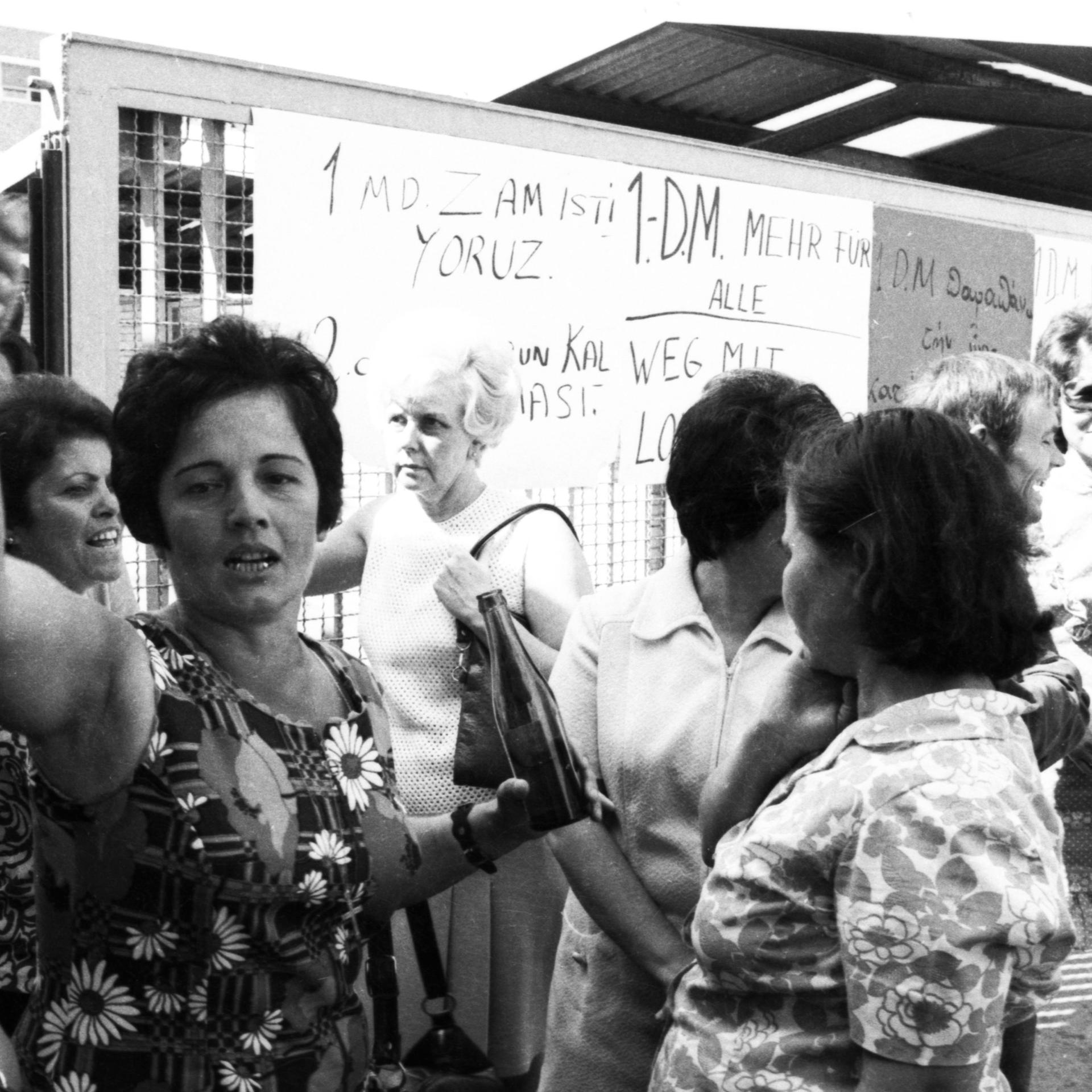 Eine Gruppe von Frauen steht vor einer Tafel auf der etwas aufgeschrieben ist. Das Bild wirkt als wäre es im Streikgetümmel entstanden.