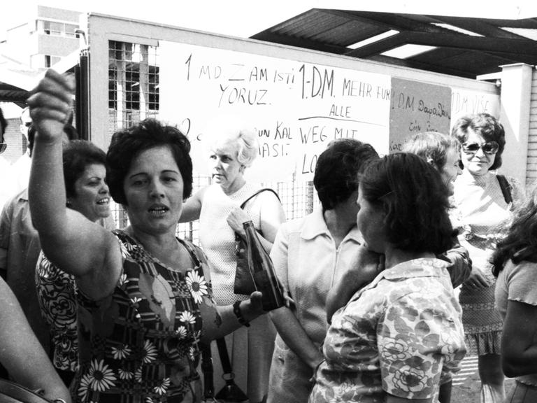Eine Gruppe von Frauen steht vor einer Tafel, auf der etwas aufgeschrieben ist. Das Bild wirkt, als wäre es im Streikgetümmel entstanden.