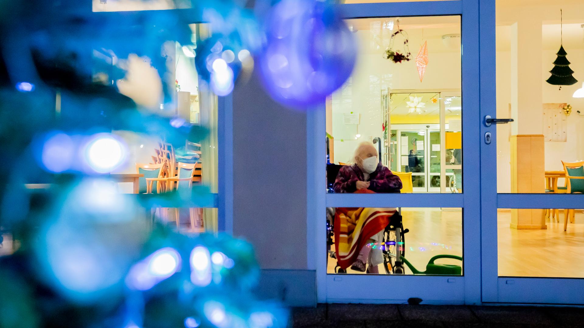 Eine Bewohnerin eines Seniorenzentrums sitzt mit Mund-Nasen-Schutz in einem Rollstuhl hinter einer Fensterscheibe. Eine Bewohnerin eines Seniorenzentrums sitzt mit Mund-Nasen-Schutz in einem Rollstuhl hinter einer Fensterscheibe.