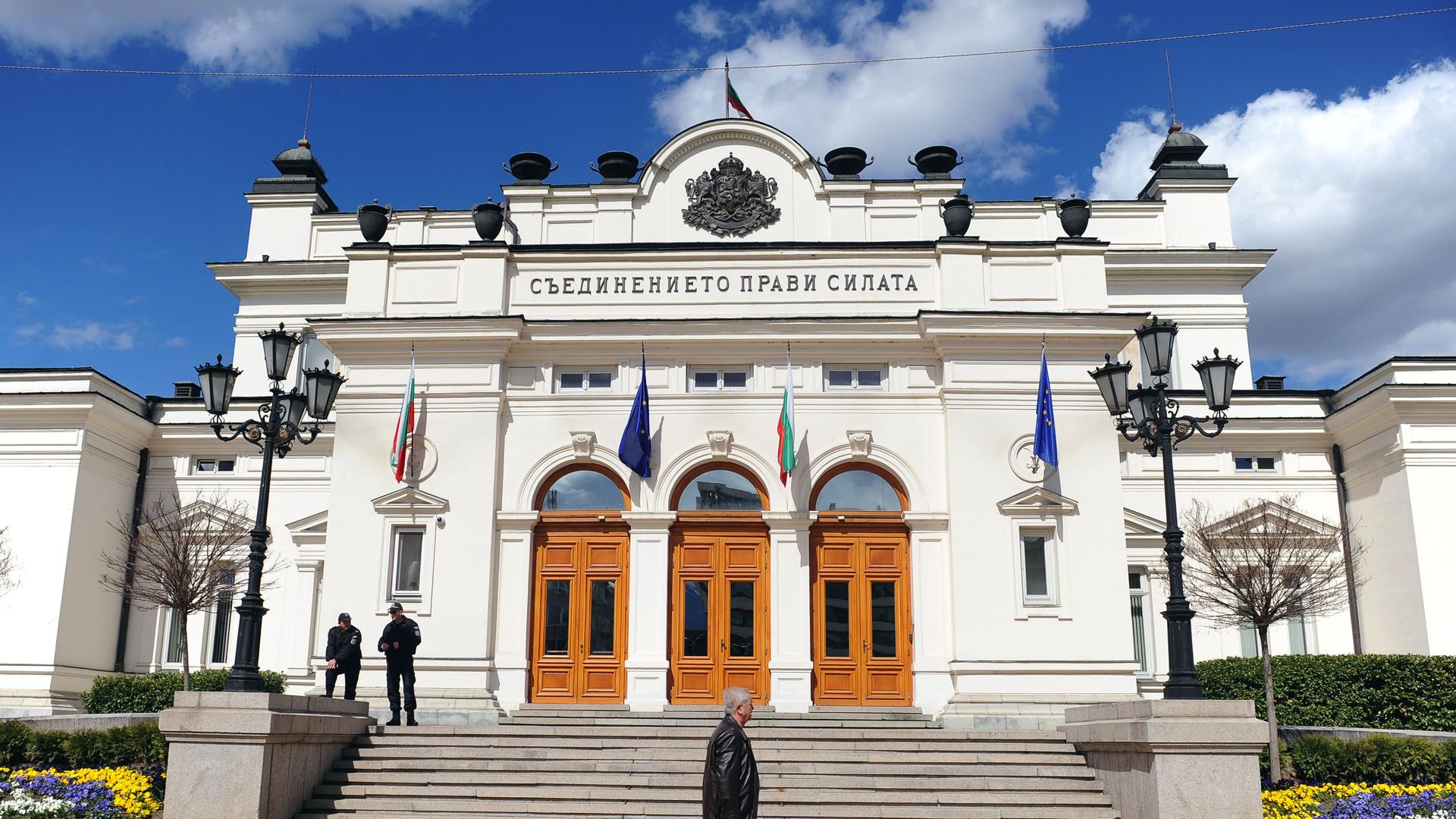 Das weiße Parlamentsgebäude hat drei braune Eingangstüren und ist mit Flaggen geschmückt. Mehrere Stufen führen zum Eingang. Das weiße Parlamentsgebäude hat drei braune Eingangstüren und ist mit Flaggen geschmückt. Mehrere Stufen führen zum Eingang.