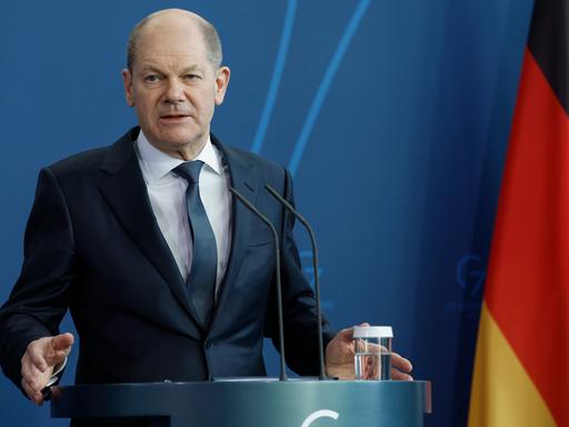 Bundeskanzler Olaf Scholz (SPD) steht an einem Rednerpult vor einer blauen Wand und einer Deutschlandfahne.