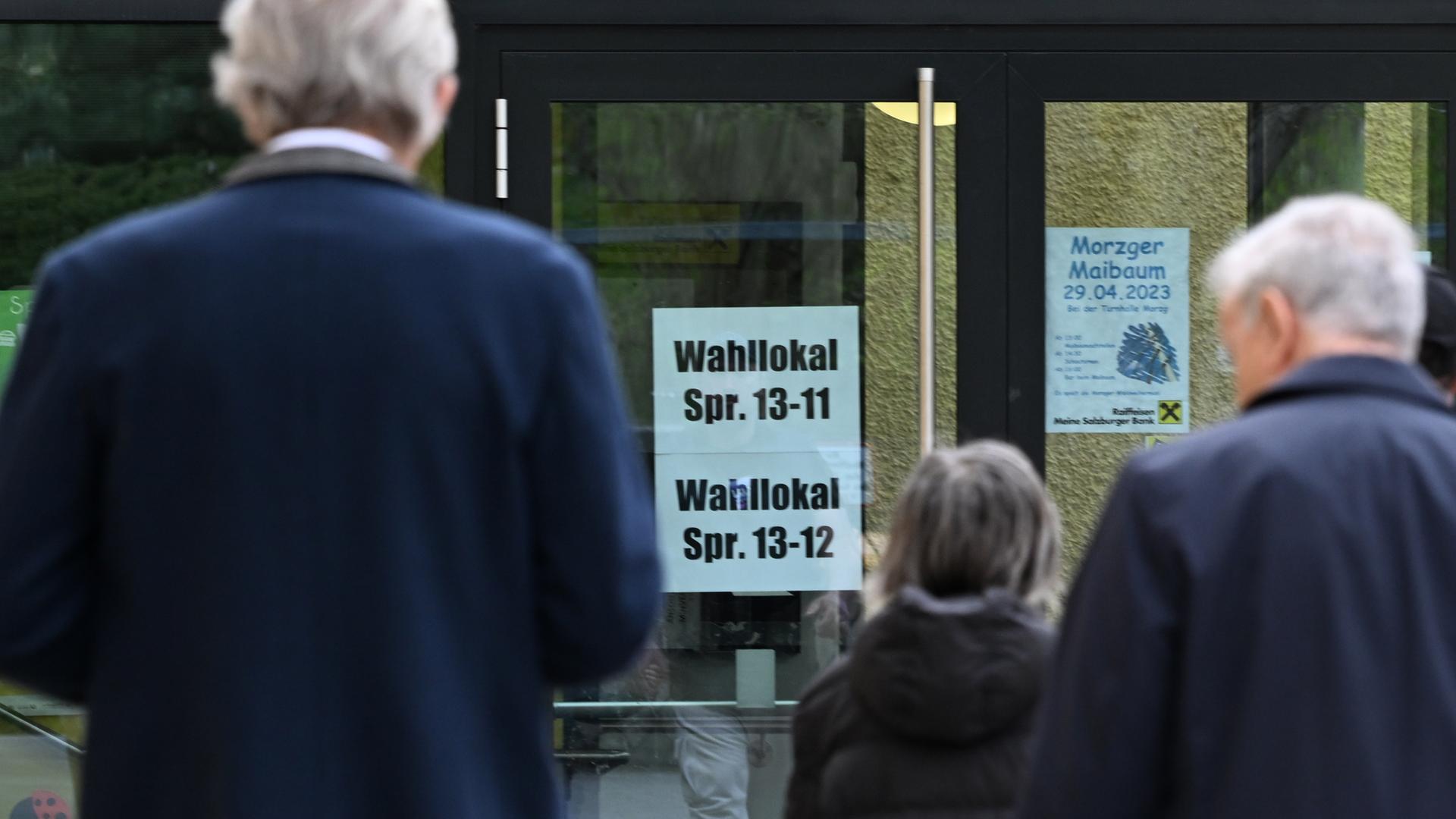 Man sieht im Vordergrund unscharf eine Frau und zwei Männer, die in Richtung des Eingangs gehen, der scharf abgebildet ist. An der Glastür kleben Schilder mit den Nummern der Stimmbezirke.