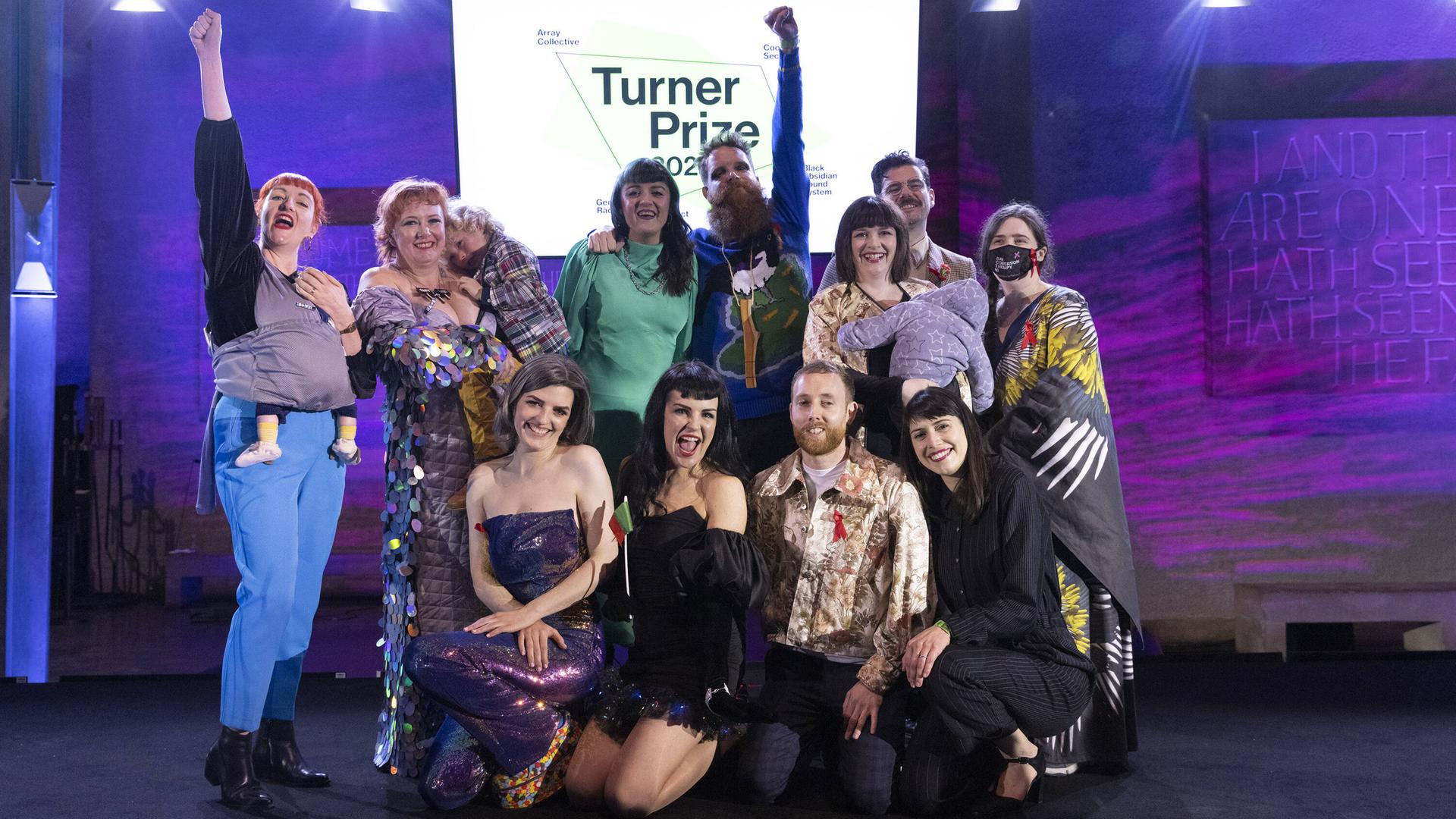 11 Menschen stehen auf einer Bühne vor einem Schild "Turner-Preis", einige recken die Faust in die Höhe, einige tragen Kinder auf dem Arm. Alle freuen sich. Es ist die Kunstgruppe Array Collective. 11 Menschen stehen auf einer Bühne vor einem Schild "Turner-Preis", einige recken die Faust in die Höhe, einige tragen Kinder auf dem Arm. Alle freuen sich. Es ist die Kunstgruppe Array Collective.