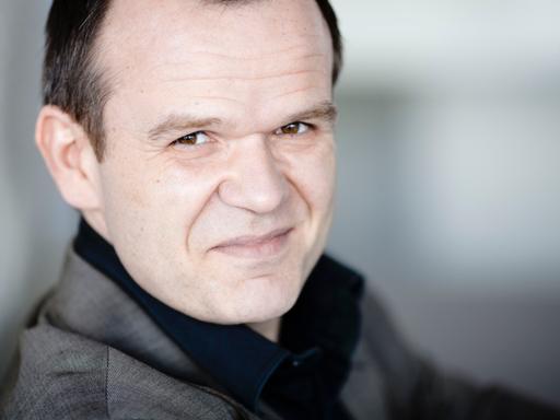 Ein Mann mit kurzen braunen Haaren blickt freundlich in die Kamera. Man sieht sein Gesicht sehr nah. Er trägt ein schwarzes Hemd und einen grauen Anzug. Es ist der Dirigent François-Xavier Roth.