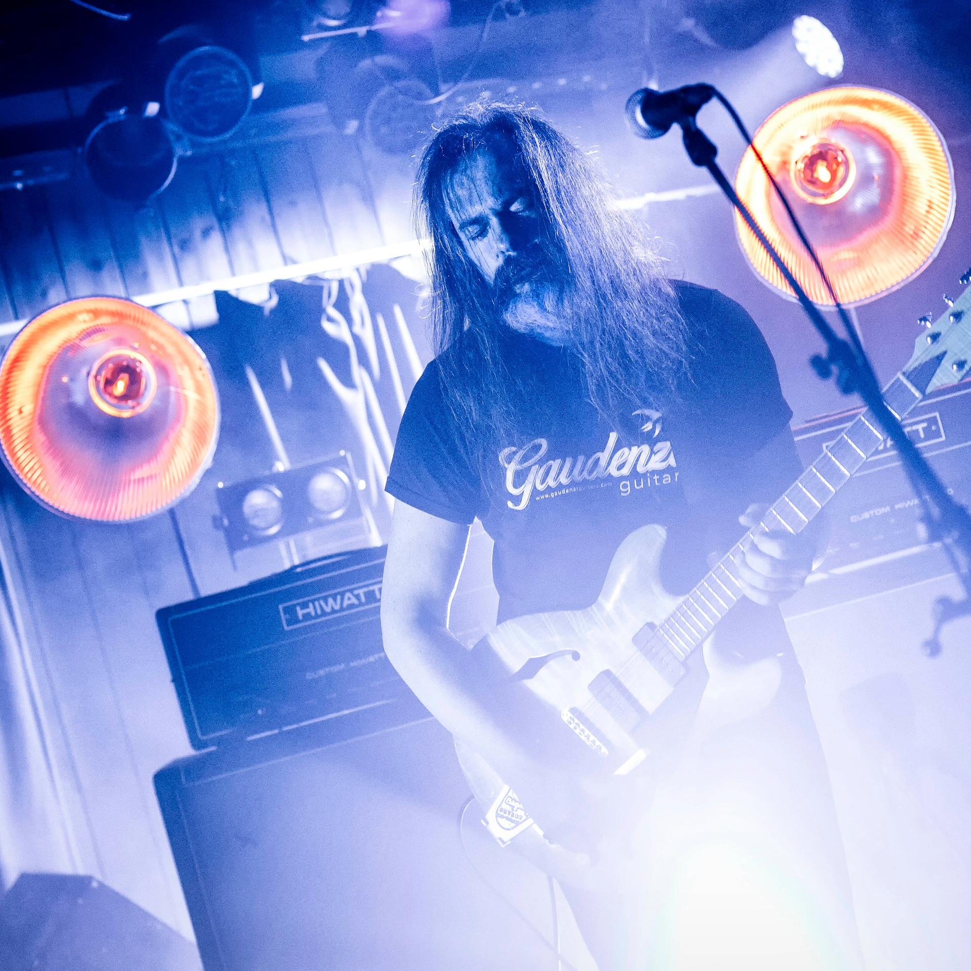 Der Sänger und Gitarrist Hans Magnus Ryan steht mit offenen langen Haaren auf einer blau beleuchteten Bühne, auf der Nebel wabert und zwei große, runde, orang-pink-farbene Scheinwerfer aufblitzen.