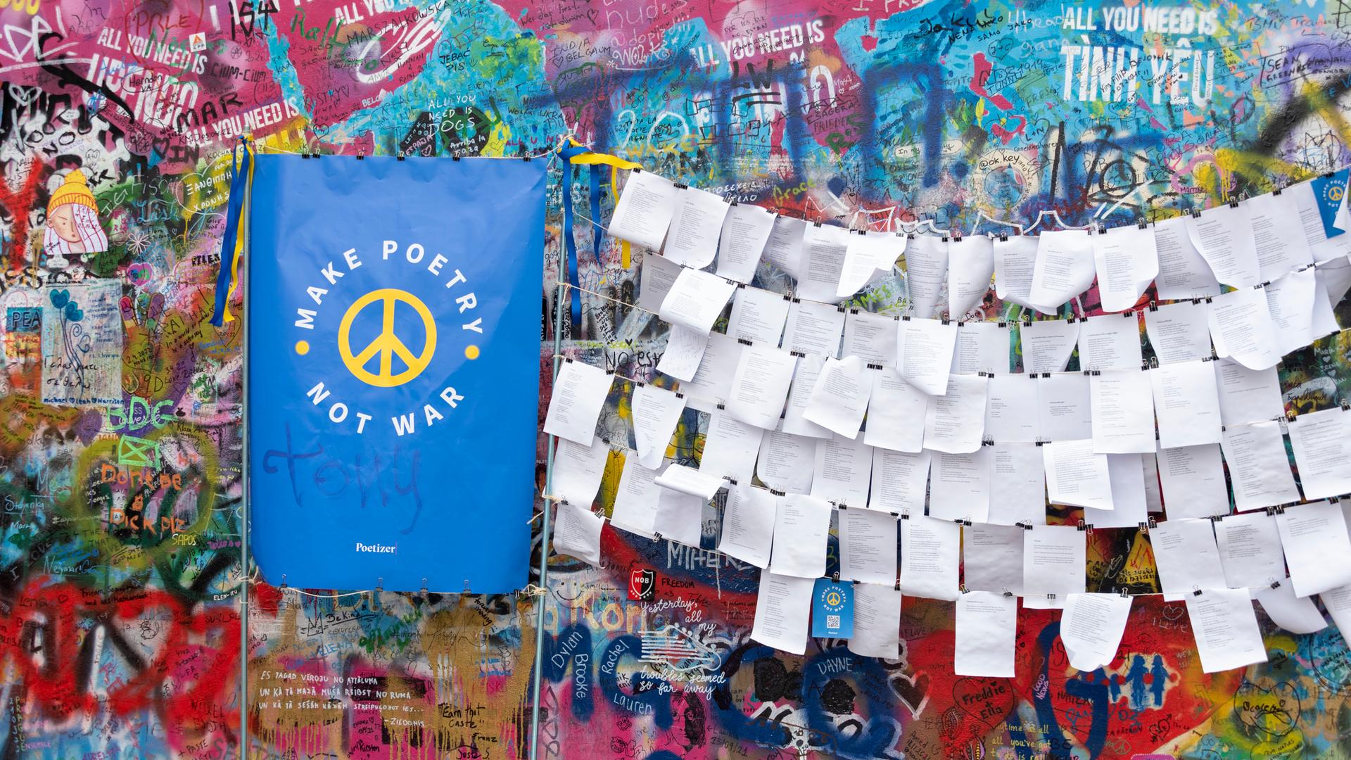 An einer mit Graffiti besprühten Mauer hängen Zettel mit Gedichten. Daneben gelehnt ein blauer Spruchbanner, auf dem in Gelb steht: "Make poetry, not war." An einer mit Graffiti besprühten Mauer hängen Zettel mit Gedichten. Daneben gelehnt ein blauer Spruchbanner, auf dem in Gelb steht: "Make poetry, not war."