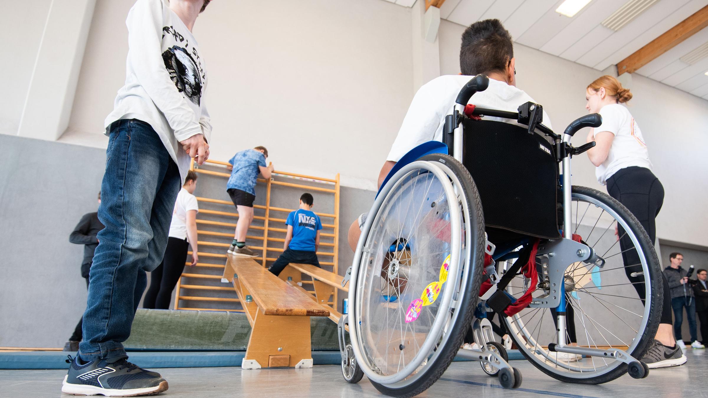 Ein Junge im Rollstuhl und Kinder ohne Behinderung beim Sportunterricht in einer Turnhalle. 