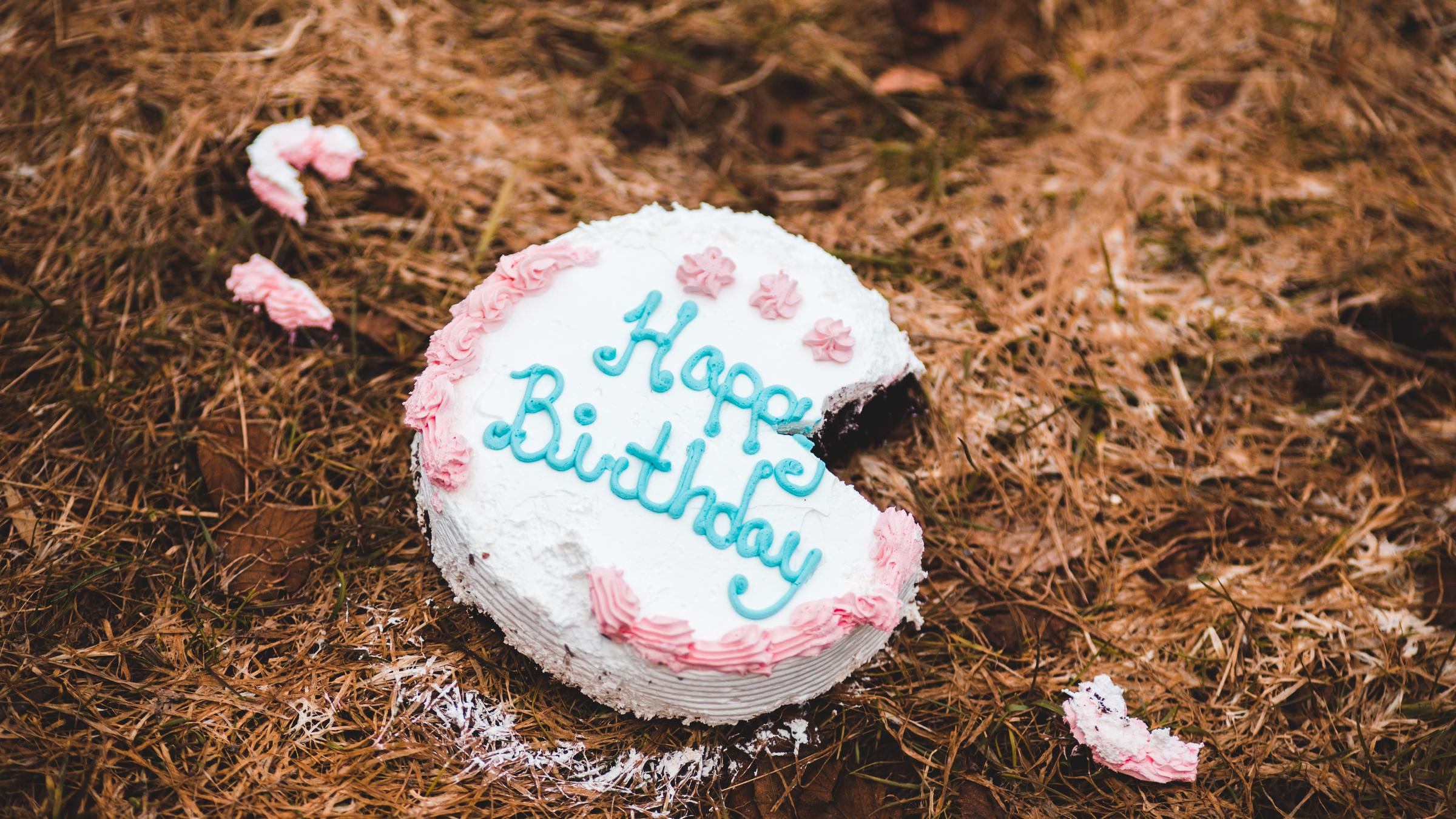 Eine Torte mit der Aufschrift Happy Birthday liegt auf dem Boden. Ein Stück fehlt und liegt in kleinen Teilen verstreut daneben. Eine Torte mit der Aufschrift Happy Birthday liegt auf dem Boden. Ein Stück fehlt und liegt in kleinen Teilen verstreut daneben.
