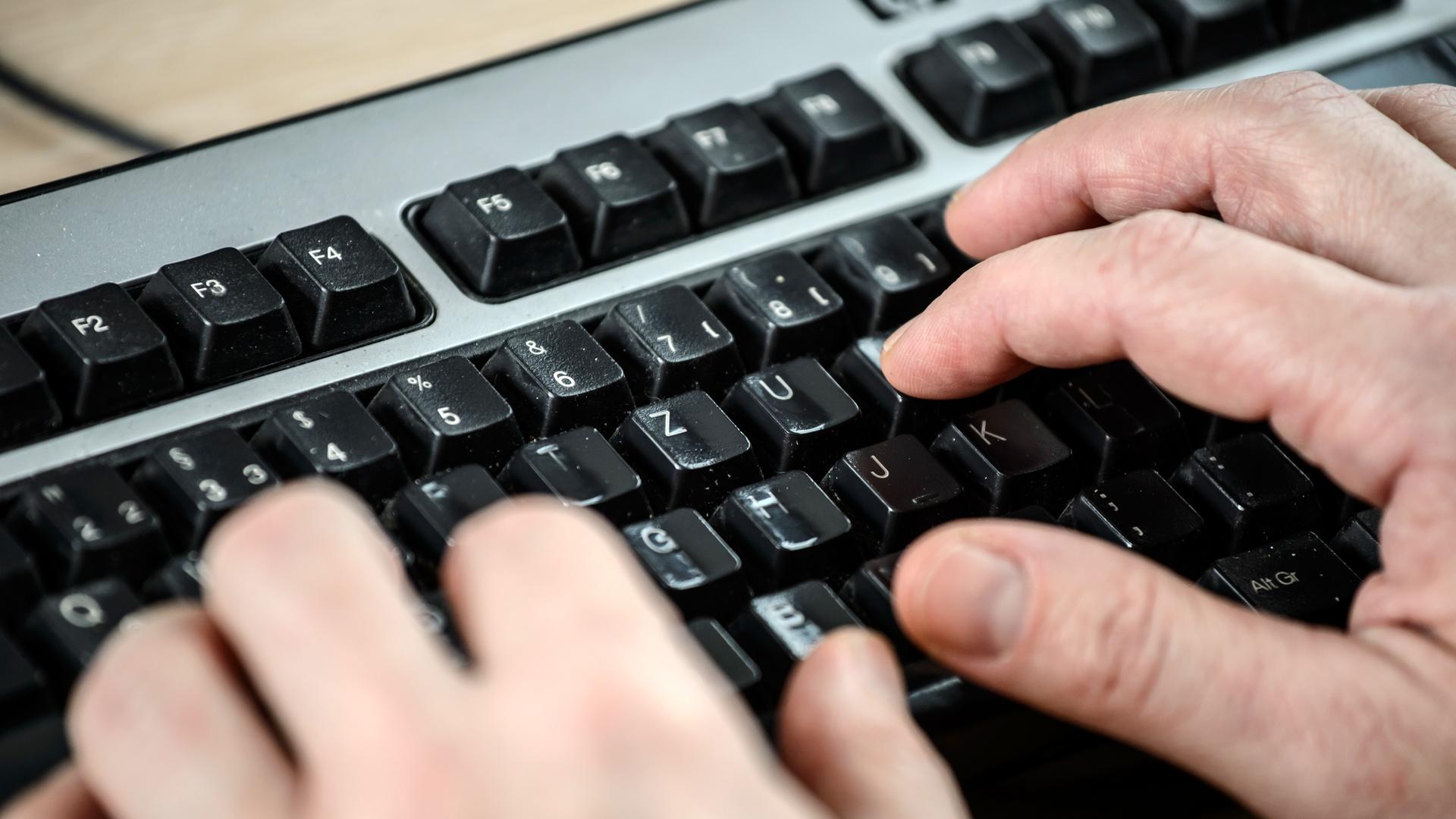Das Bild zeigt zwei Hände in einer halbnahen Aufnahme im Anschnitt. Die Finger sind über schwarzen Tasten einer grauen Computertastatur zu sehen.